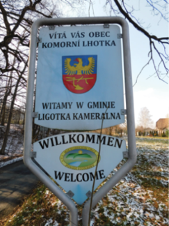 Dwa zdjęcia obok siebie. Na pierwszym zdjęciu jest krajobraz z drogą, skrzyżowaniem o ruchu okrężnym oraz ekranami akustycznymi drogi szybkiego ruchu w tle. Na pierwszy plan wysuwają się dwie tablice drogowe „koniec gminy Bystřice” w języku czeskim oraz „koniec gminy Bystrzyca“ w języku polskim. Czeską tablicę zainstalowano na metalowym słupku wyżej od polskiej. Wielkością, kształtem, kolorami i czcionką są obydwie identyczne. Na drugim zdjęciu zobaczyć można „tablicę powitalną“, zainstalowaną przy drodze w otoczeniu drzew. W środku metalowej tablicy w kształcie prostokątu i niewielkiego trójkątu widnieje znak gminy Ligotka Kameralna (Komorní Lhotka), wokół którego zamieszczono napisy w języku czeskim, polskim, niemieckim i angielskim „Vítá vás obec Komorní Lhotka / Witamy w gminie Ligotka Kameralna / Willkommen / Welcome.