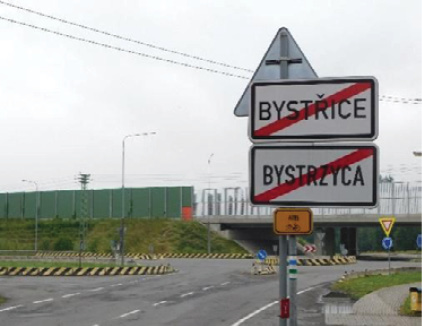 Dwa zdjęcia obok siebie. Na pierwszym zdjęciu jest krajobraz z drogą, skrzyżowaniem o ruchu okrężnym oraz ekranami akustycznymi drogi szybkiego ruchu w tle. Na pierwszy plan wysuwają się dwie tablice drogowe „koniec gminy Bystřice” w języku czeskim oraz „koniec gminy Bystrzyca“ w języku polskim. Czeską tablicę zainstalowano na metalowym słupku wyżej od polskiej. Wielkością, kształtem, kolorami i czcionką są obydwie identyczne. Na drugim zdjęciu zobaczyć można „tablicę powitalną“, zainstalowaną przy drodze w otoczeniu drzew. W środku metalowej tablicy w kształcie prostokątu i niewielkiego trójkątu widnieje znak gminy Ligotka Kameralna (Komorní Lhotka), wokół którego zamieszczono napisy w języku czeskim, polskim, niemieckim i angielskim „Vítá vás obec Komorní Lhotka / Witamy w gminie Ligotka Kameralna / Willkommen / Welcome.