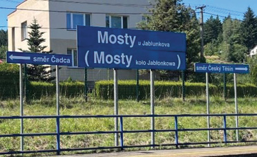 [Dwa zdjęcia obok siebie. Na pierwszym zdjęciu znajduje się fragment peronu dworca kolejowego. W tle drzewa, dom jednorodzinny, trawnik. Przy peronie jest kilka metalowych słupków, na których znajduje się metalowe tablice – niebieskie prostokąty z białą czcionką. W środku na tablicy zamieszczonej najwyżej jest napis „Mosty u Jablunkova”, poniżej jest tablica z napisem w nawiasie „(Mosty koło Jabłonkowa)”. Wlewo jest mniejsza tablica ze strzałką wskazującą wlewo i napisem „směr Čadca”, wprawo jest taka sama tablica ze strzałką wskazującą wprawo i napisem „směr Český Těšín”. Oprócz nawiasu zastosowanego w polskim napisie nazwy stacji, są formlanie wszystkie napisy tożsame.]
